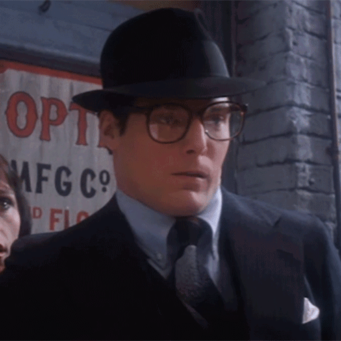A Man Wearing Glasses And A Hat Is Standing In Front Of A Sign That Says Opti Mfg Co GIF