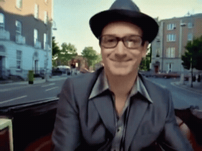A Man Wearing Glasses And A Hat Smiles While Sitting In A Car GIF