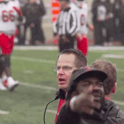 A Man Wearing Glasses And A Hat Stands On A Football Field In Front Of A Referee With The Number 66 On His Jersey GIF