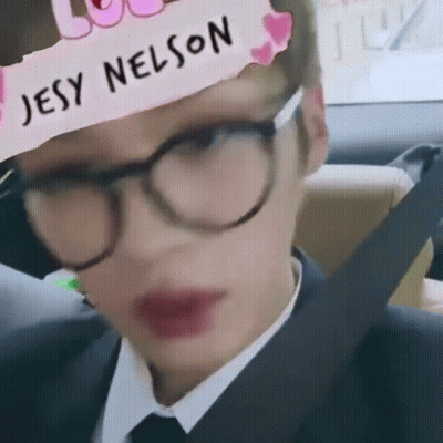 A Man Wearing Glasses And A Hat With The Name Jesy Nelson On It Is Sitting In A Car . GIF