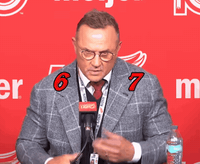 A Man Wearing Glasses And A Lanyard With The Number 6 And 7 On It Stands In Front Of A Red Background GIF