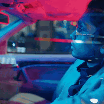 A Man Wearing Glasses And A Necklace Is Driving A Car At Night GIF