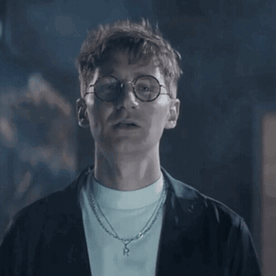 A Man Wearing Glasses And A Necklace Is Looking At The Camera In A Dark Room . GIF