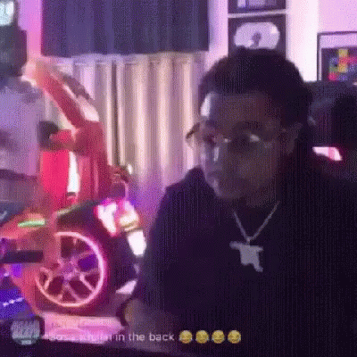 A Man Wearing Glasses And A Necklace Is Sitting In Front Of A Car . GIF