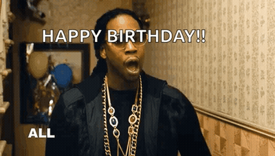 A Man Wearing Glasses And A Necklace Is Standing In A Hallway . GIF