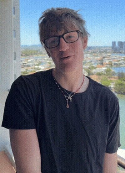 A Man Wearing Glasses And A Necklace Stands In Front Of A View Of A City GIF