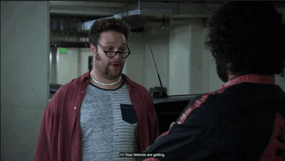 A Man Wearing Glasses And A Necklace Talks To Another Man In A Parking Garage GIF