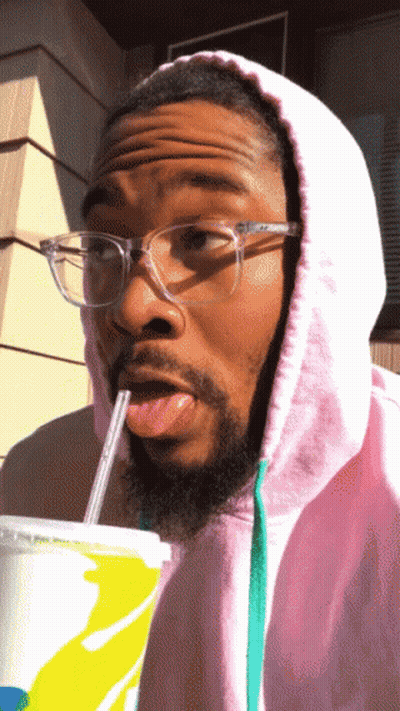 A Man Wearing Glasses And A Pink Hoodie Drinks From A Cup With A Straw GIF
