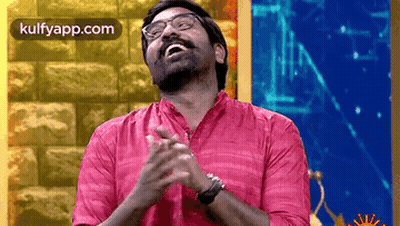 A Man Wearing Glasses And A Pink Shirt Is Clapping His Hands In Front Of A Brick Wall . GIF