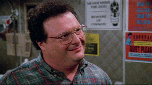 A Man Wearing Glasses And A Plaid Shirt Smiles In Front Of A Sign That Says " Never Mind The Dog " GIF