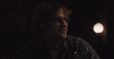 A Man Wearing Glasses And A Plaid Shirt Smiles In The Dark GIF