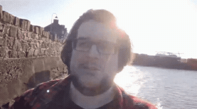 A Man Wearing Glasses And A Plaid Shirt Stands In Front Of A Body Of Water GIF