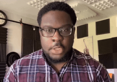 A Man Wearing Glasses And A Plaid Shirt Stands In Front Of A Wall With A Clock On It GIF