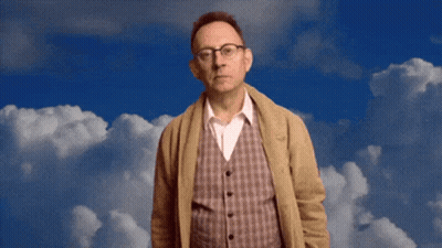 A Man Wearing Glasses And A Plaid Vest Stands In Front Of A Blue Sky With White Clouds GIF