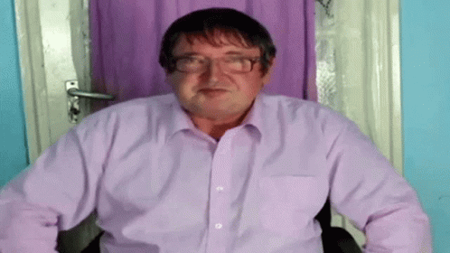 A Man Wearing Glasses And A Purple Shirt Is Sitting In A Chair . GIF