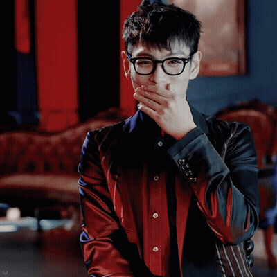A Man Wearing Glasses And A Red And Black Jacket Is Covering His Mouth With His Hand GIF
