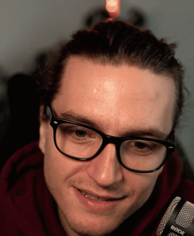 A Man Wearing Glasses And A Red Hoodie Is Smiling In Front Of A Rode Microphone GIF
