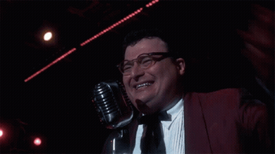 A Man Wearing Glasses And A Red Jacket Is Smiling In Front Of A Microphone GIF