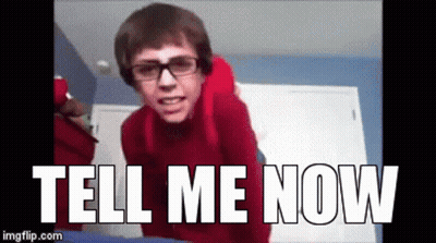 A Man Wearing Glasses And A Red Sweater Is Standing In Front Of A Sign That Says Tell Me Now GIF