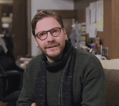 A Man Wearing Glasses And A Scarf Is Sitting On A Couch GIF
