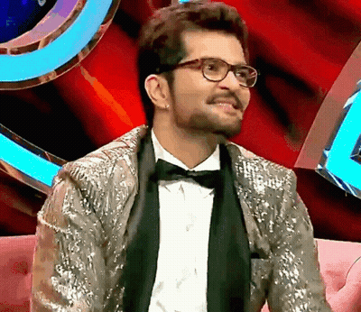 A Man Wearing Glasses And A Sequined Jacket Is Sitting On A Couch GIF