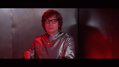 A Man Wearing Glasses And A Silver Shirt Is Pointing At The Camera GIF