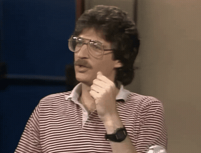 A Man Wearing Glasses And A Striped Shirt Holds His Hand To His Chin GIF