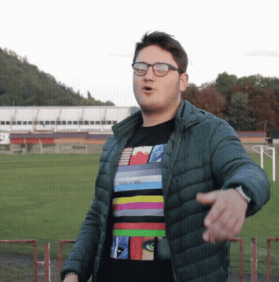 A Man Wearing Glasses And A Striped Shirt Stands In Front Of A Soccer Field GIF