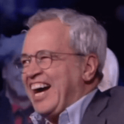 A Man Wearing Glasses And A Suit Is Laughing In A Crowd . GIF