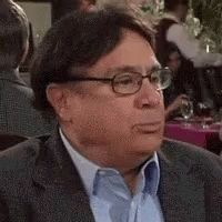 A Man Wearing Glasses And A Suit Is Sitting At A Table With Other People . GIF