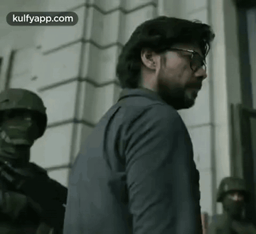 A Man Wearing Glasses And A Suit Is Standing In Front Of A Building With Soldiers . GIF