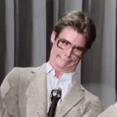 A Man Wearing Glasses And A Suit Is Standing In Front Of A Microphone Making A Funny Face . GIF