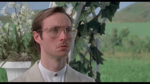 A Man Wearing Glasses And A Suit Stands In Front Of A Flower Pot GIF