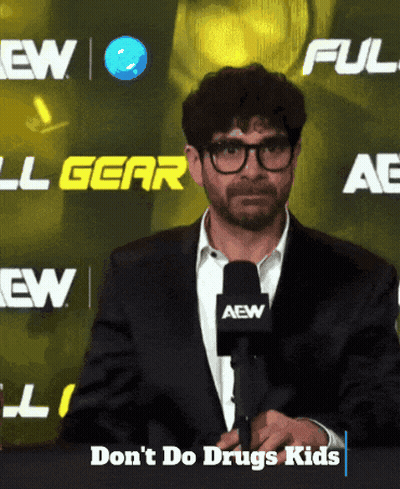 A Man Wearing Glasses And A Suit Stands In Front Of A Sign That Says Aew GIF