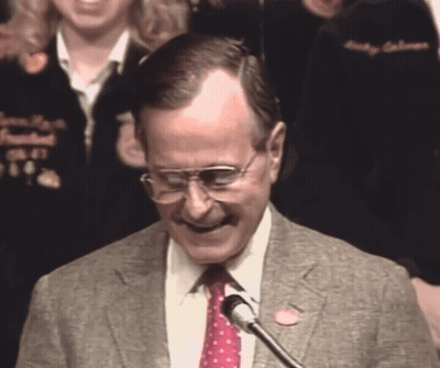 A Man Wearing Glasses And A Tie Is Smiling At A Microphone GIF