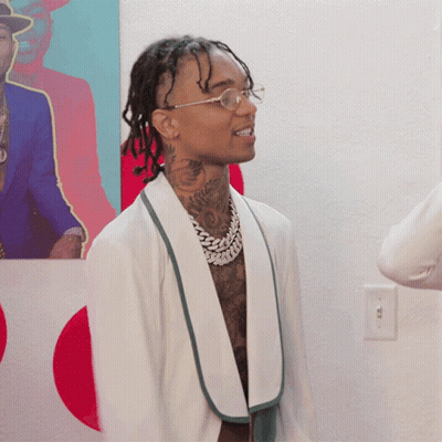 A Man Wearing Glasses And A White Jacket Stands In Front Of A Painting GIF