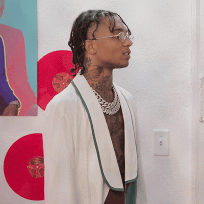 A Man Wearing Glasses And A White Robe Is Standing In Front Of A Wall With Red Records On It GIF