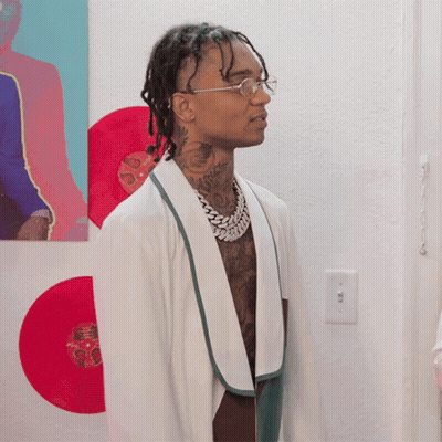 A Man Wearing Glasses And A White Robe Stands In Front Of A Wall With Red Records GIF