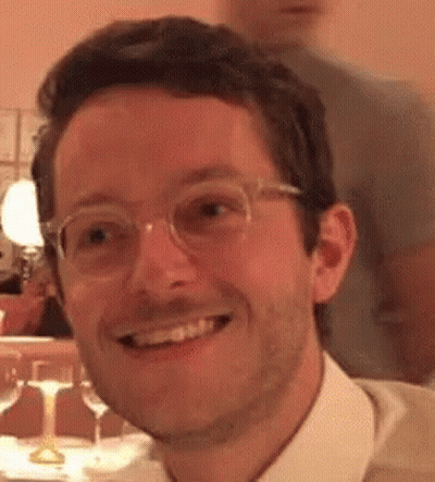 A Man Wearing Glasses And A White Shirt Is Smiling And Looking At The Camera . GIF