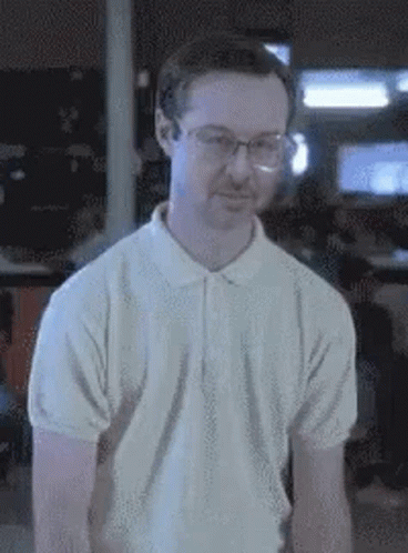 A Man Wearing Glasses And A White Shirt Is Standing In A Dark Room . GIF