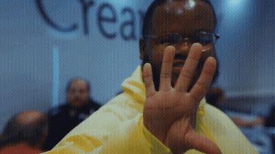 A Man Wearing Glasses And A Yellow Jacket Holds Up His Hand In Front Of A Wall That Says Crea GIF