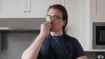 A Man Wearing Glasses And An Apron Smells Something In A Kitchen GIF