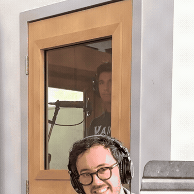 A Man Wearing Glasses And Headphones Stands In Front Of A Door That Has A Window GIF