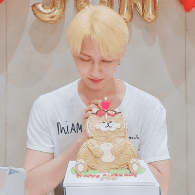 A Man Wearing Glasses Blows Out A Pink Candle On Top Of A Cat Cake GIF