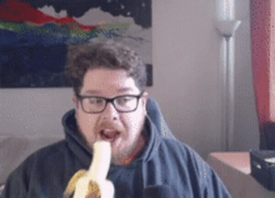 A Man Wearing Glasses Is Eating A Banana In Front Of A Painting GIF