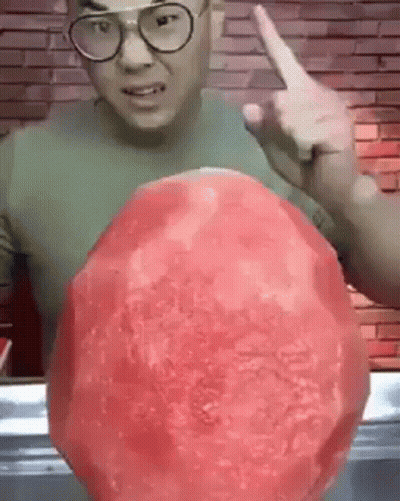 A Man Wearing Glasses Is Holding A Large Watermelon In His Hand And Pointing At It . GIF