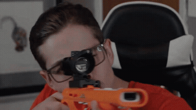 A Man Wearing Glasses Is Holding A Nerf Gun In Front Of His Face GIF