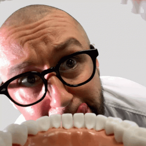 A Man Wearing Glasses Is Licking A Model Of His Teeth GIF