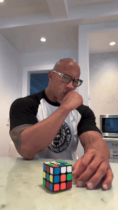 A Man Wearing Glasses Is Playing With A Rubik 'S Cube On A Countertop GIF
