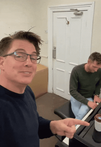 A Man Wearing Glasses Is Pointing To Another Man Playing A Piano GIF